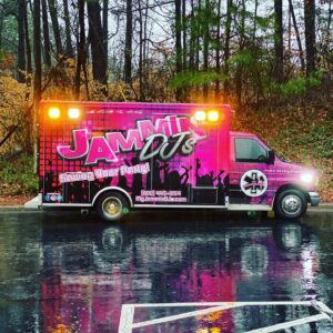 A vibrant pink and black jammin' djs van parked on a wet parking lot, surrounded by trees, with its lights on during rainfall.