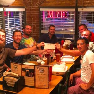 Group of seven men smiling and toasting at a pub trivia night, illuminated by warm light with neon signs visible in the background.