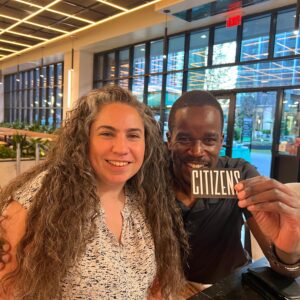 A man and a woman sitting at a table indoors, smiling at the camera; the man is holding up a card that says 
