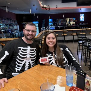 Two people in skeleton costumes sit at a restaurant table. They are smiling, and a red card is held by one of them. Glasses and condiments are on the table.