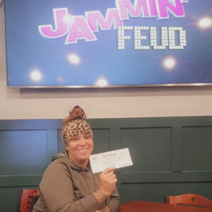 A woman sitting at a table holds up a check and smiles in front of a screen displaying the words 