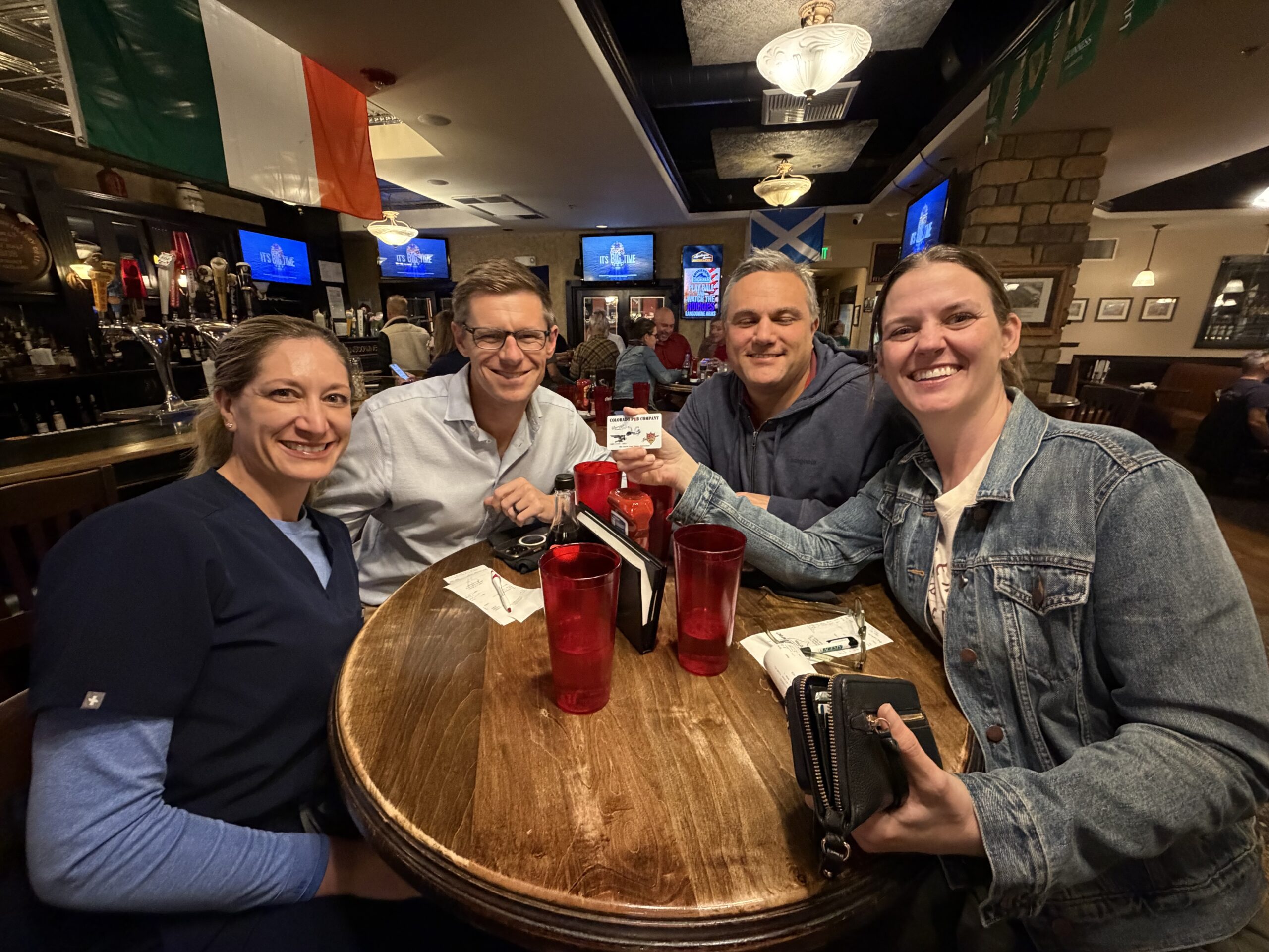 Four people sit around a wooden table in a pub, smiling at the camera. There are drinks and receipts on the table, and an Irish flag hangs in the background.