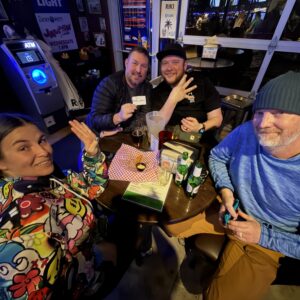 Four adults sit around a bar table with drinks, snacks, and game cards, smiling and posing for the camera in a lively, colorful setting.