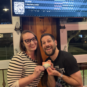 Two people smiling and holding a gift card in front of a screen displaying trivia results at a bar or restaurant.