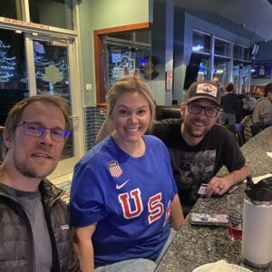 Three adults sit at a bar counter smiling at the camera. One wears a blue U.S. sports shirt. Drinks, menus, and a water bottle are on the counter; TVs and people are visible in the background.